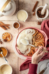 Female hands pouring glaze over freshly baked cinnabon buns in ceramic baking dish, beige tile background. Making cinnamon rolls process, top view