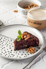 Chocolate spongy brownie cake served with walnuts and mint leaves on top and coffee on white tile background. A piece of chocolate brownie cake