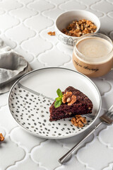 Chocolate spongy brownie cake served with walnuts and mint leaves on top and coffee on white tile background. A piece of chocolate brownie cake