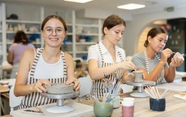 Three girls craftsperson are busy doing vase from clay and painting ceramic products. They sitting near desktop, using tools, busy with hobbies leisure activity. Working together in pottery workshop.