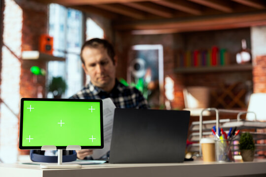 Green screen tablet in front of man in home office crosschecking information between laptop screen and paper files. Close up of chroma key device next to employee checking documents - Powered by Adobe