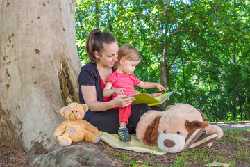 Mother reading a book with her cute little child under an old plane tree in the park. Funny and educative outdoor activity for children, in sunny summer day