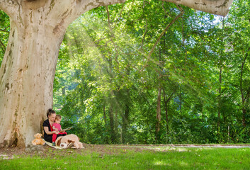 Mother reading a book with her cute little child under an old plane tree in the park. Funny and educative outdoor activity for children, in sunny summer day
