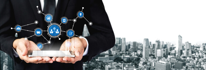 A businessman holds a tablet displaying digital icons symbolizing technology and networking, set against a modern city skyline, representing innovation and success. Tessel