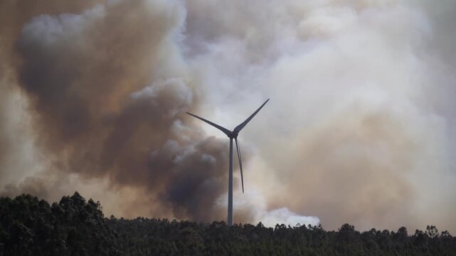 A dramatic wildfire burns near a windmill under a sky filled with thick clouds. The contrast between renewable energy and destructive flames highlights themes of climate change, environmental crisis