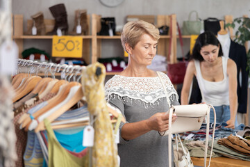 Mature woman stands in a store in the sale area of clothes and accessories and carefully chooses a...