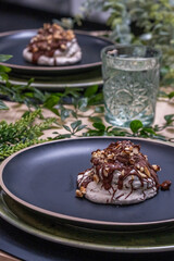 Meringue dessert topped with rich chocolate and crunchy nuts, elegantly presented on a dark plate surrounded by greenery and a refreshing glass of water with copy space