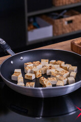 Cubes of golden-brown tofu are sizzling in a non-stick frying pan on a modern stovetop, showcasing the cooking process and vibrant kitchen environment
