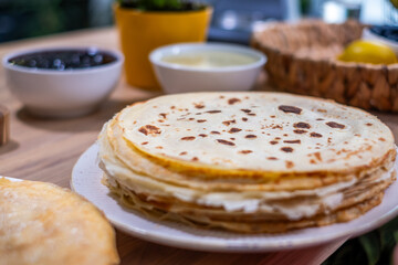 Stack of freshly made crepes on a white plate, surrounded by bowls of jam and cream, with a vibrant plant in the background, showcasing delicious breakfast options