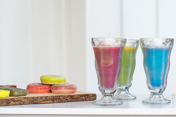 Pink, green and blue colored wax in tall slender parfait glasses on a white display table with a light  background. A tray of round soap bars; green, yellow and orange color sits on the table.