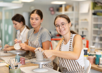 Adolescence girl student sits near desktop and doing from clay product. Lover of applied art completes design of bowl made of clay, forms final appearance of product