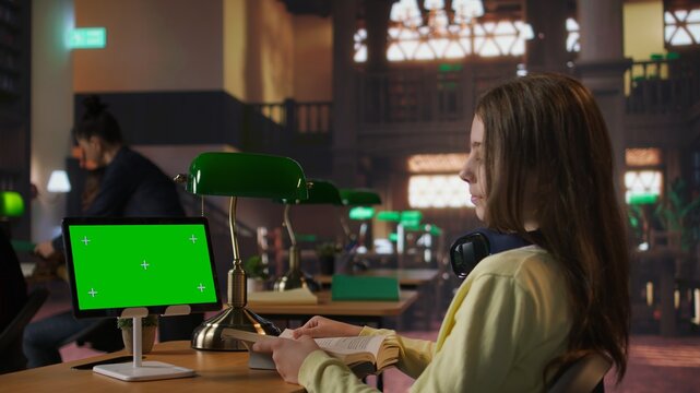 Primary school student sitting at desk in the library and reading next to mockup on tablet, preparing notes and ideas for future lessons. Scholar girl using digital gadget for assignments. Camera A.