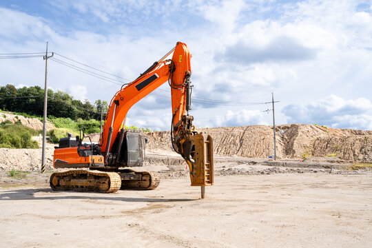 Yellow excavator with a hydraulic breaker attachment working at a construction site on a sunny day. Heavy machinery used for demolition, rock breaking, and ground preparation. Suitable for constructio