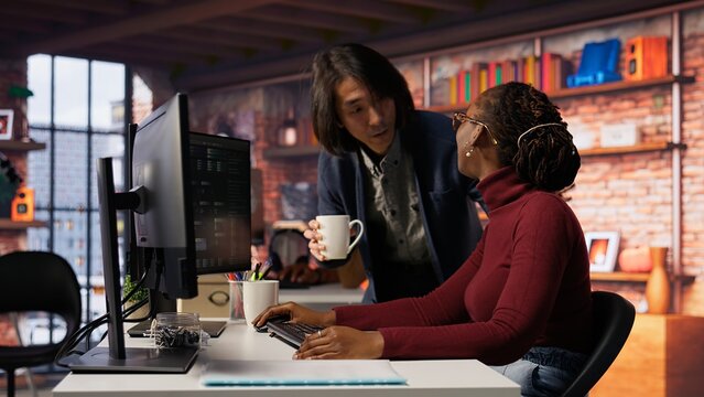 Exhausted AI startup employee rubbing forehead while analyzing datasets on computer, receiving coffee from project CTO manager to get energy and continue coding, camera B