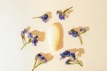 Yellow cosmetic tube surrounded by dried blue flowers on light background. Top view, flat lay, mockup