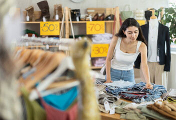 Young woman customer making choice and elect pants in store, and buyer consider some items. Modern urban fashion, casual practical clothes made of natural fabrics