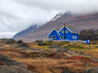 Bright blue house in wild autumn terrain with volcanic rocks and fog-covered mountains in the background.