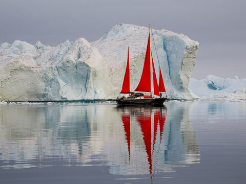 Red-sailed boat sailing through icy arctic waters surrounded by floating ice and large icebergs under cloudy sky.