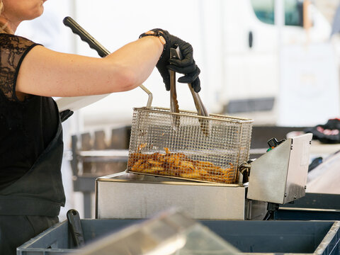 Pr&eacute;paration de frites dor&eacute;es dans une friteuse professionnelle en acier inoxydable, sc&egrave;ne de cuisine de rue ou de f&ecirc;te avec vendeuse au travail.