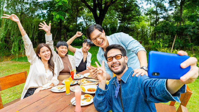 Group of Happy Asian people diverse friends using mobile phone taking selfie together while celebration social gathering meeting dinner party eating food, drinking cocktail in the garden in evening.