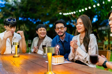 Group of Happy Diverse Asian millennial people friends enjoy celebration birthday party having dinner together in the garden. Beautiful woman blowing candle on birthday cake on dining table.