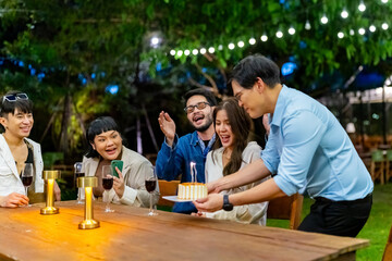 Group of Happy Diverse Asian millennial people friends enjoy celebration birthday party having dinner together in the garden. Beautiful woman blowing candle on birthday cake on dining table.