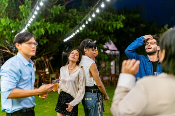 Fototapeta premium Happy diverse Asian millennial people friends enjoy and fun meeting holiday celebration social gathering dinner party in garden evening, drinking beer and dancing together outdoor festive event.
