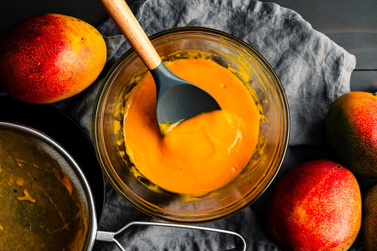 Freshly Made mango Puree in a Glass Mixing Bowl: Directly above strained mango pulp with a mesh strainer and fresh mangoes on the side