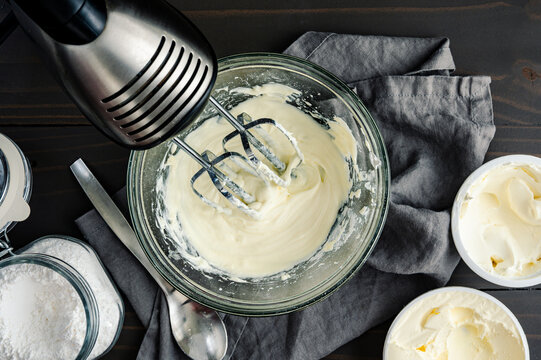 Homemade Cream Cheese Frosting with hand Mixer: Glass mixing bowl of beaten heavy whipping cream, mascarpone cream, and powdered sugar 