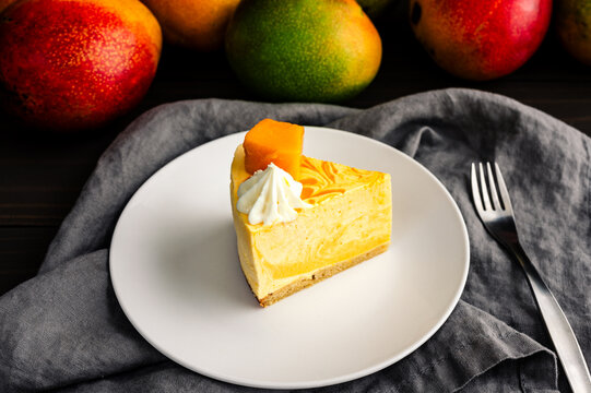 Slice of Mango Mousse Cake on a Plate with Fresh Mangoes: Angled view of a piece of swirled mousse cake with cream cheese frosting on a dessert plate