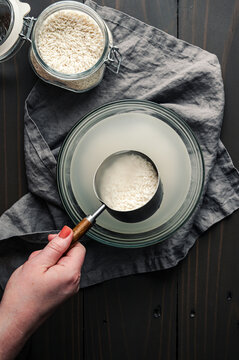 Hand Holding a Measuring Cup of Soaking Sticky Rice: Scoop of glutinous rice held over a glass bowl full of cloudy water and soaked sweet rice