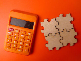 The image displays a small orange electronic calculator alongside four interlocking wooden puzzle pieces on an orange background