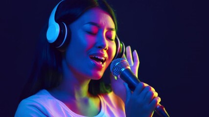 A young Asian woman sings into a microphone while wearing headphones, surrounded by colorful neon lights. The energetic atmosphere captures a moment of artistic expression and joy. - Powered by Adobe