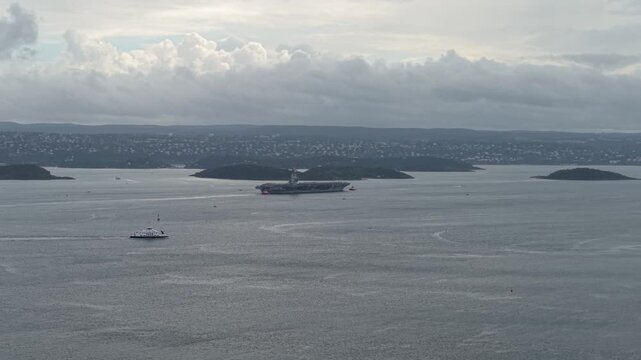 Boat Cruising In The Fjord Passing By The USS Gerald R. Ford Aircraft Carrier Ship In Oslo, Norway. - aerial shot