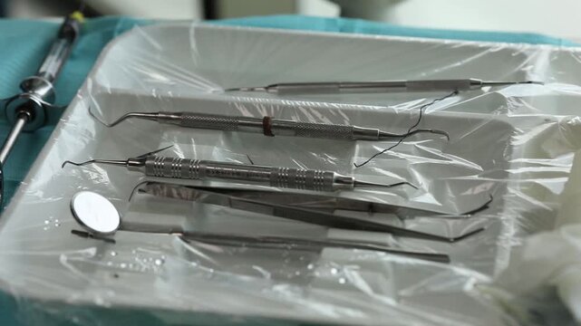 Sterile dental tray covered with film holding mirror, tweezers, two probes, anesthesia syringe, and retraction cord. Instruments prepared for dental treatment.
