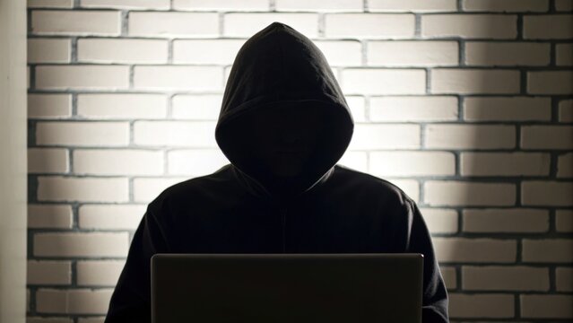 A shadowy figure in a hoodie sits before a laptop, silhouetted against a brick wall, evoking themes of mystery and digital intrigue.