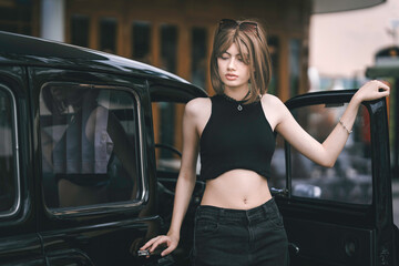 Beautiful young woman in a stylish black outfit stepping into a vintage black car outdoors. The fashionable portrait of a pretty teenager in a retro style.