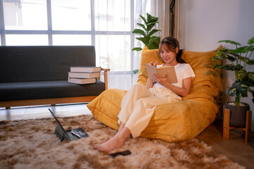 Obraz premium Young woman relaxing on beanbag chair reading and taking notes in cozy living room