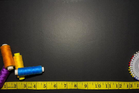 A sewing and tailoring flat lay with colorful thread spools, a measuring tape, and sewing pins on a dark background with ample copy space