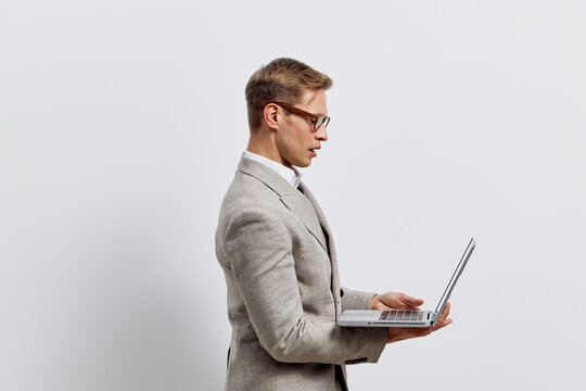 Professional young man in smart casual attire using a laptop computer while standing indoors against a plain white background. Business, technology, office, work, concentration, communication. - Powered by Adobe