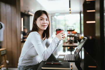 A young Asian woman sits in a coffee shop with her laptop, checking official documents, and smiling...