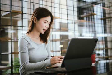 A young Asian woman sits in a coffee shop with her laptop, checking official documents, and smiling...