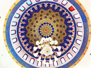 A beautifully decorated mosque dome with intricate floral and geometric patterns, illuminated by natural light from small windows around the edge.