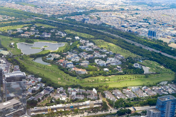 Fototapeta premium Aerial view of a large green golf course surrounded by residential areas and city buildings, showing a contrast between urban life and natural landscape with lakes, roads, and trees.