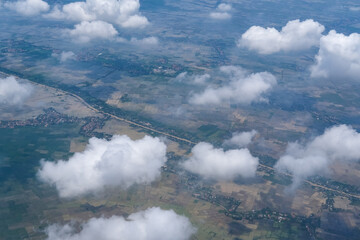 Obraz premium Aerial view of green landscapes partially covered by fluffy white clouds reveals rural towns, rice fields, and winding roads, showcasing Indonesia’s natural beauty from above the sky.