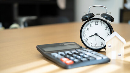Black calculator house model and alarm clock placed together on wooden table representing mortgage planning home loan repayment financial investment and time control.