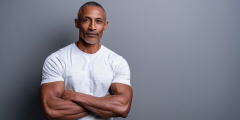 Mature personal trainer with a confident expression, standing in front of a gray backdrop, wearing a white t-shirt. 