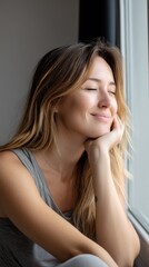 Smiling woman with peaceful expression, dreaming while sitting on a windowsill.