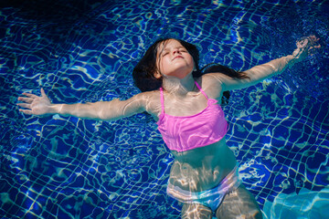 Joyful little girl in a pink swimsuit floats with arms outstretched in a swimming pool, eyes closed and smiling while enjoying the summer sun.