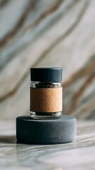 A small glass jar with a cork band and black lid sits on a dark grey stone pedestal against a marbled background